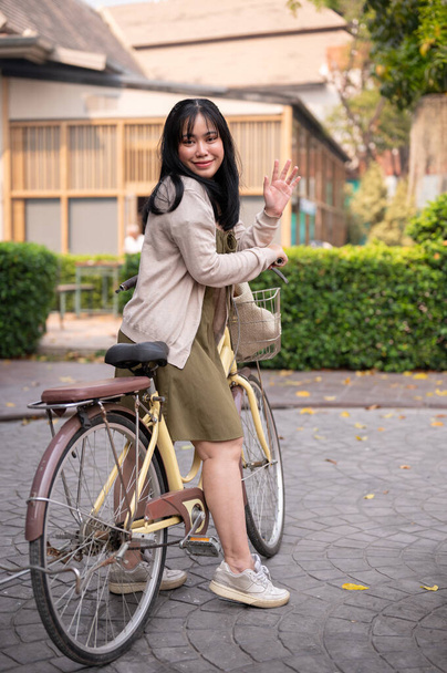Houkutteleva, hymyilevä nuori aasialainen nainen heiluttaa kättään tervehtiäkseen jotakuta pyöräillessään kaupungissa. ympäristöystävällisen elämäntavan käsite - Valokuva, kuva