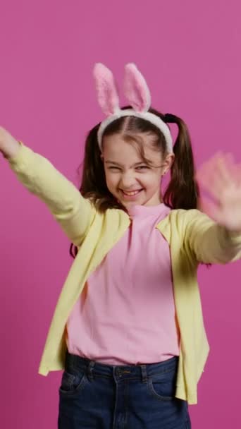 Schattig schattig kind zet konijnenoren en zwaaien naar de camera, genieten van Pasen zondag feest tegen roze achtergrond. Lachend vrolijk schoolmeisje met staartjes die hallo zeggen. Camera B. - Video
