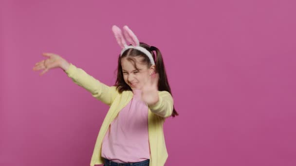 Adorable enfant mignon mettant des oreilles de lapin et agitant à la caméra, profitant Pâques dimanche célébration sur fond rose. Écolière souriante joyeuse avec des nattes disant bonjour. Caméra A. - Séquence, vidéo