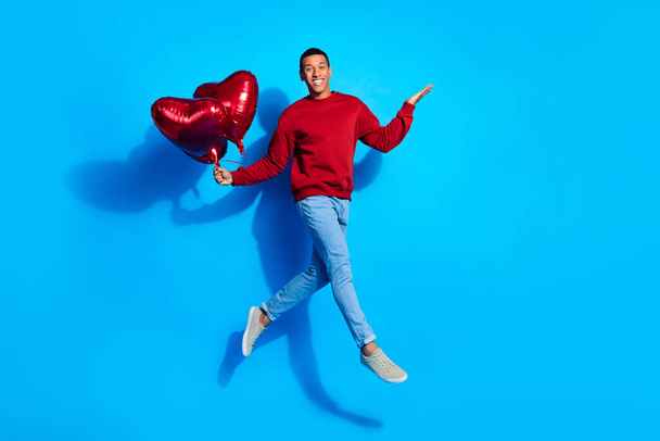 Full size photo of excited young man jump hand hold heart shape balloons empty space isolated on blue color background. - Photo, image