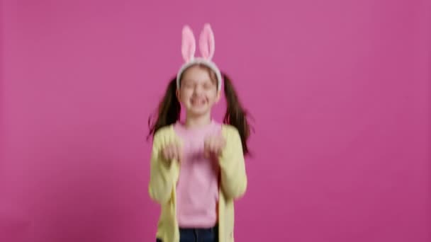 Vreugdevol zorgeloos schoolmeisje dat rondspringt in de studio, een konijn imiteert en tegen een roze achtergrond springt. Vrolijk actief kind met konijnenoren en stuiterend, schattig kind. Camera B. - Video