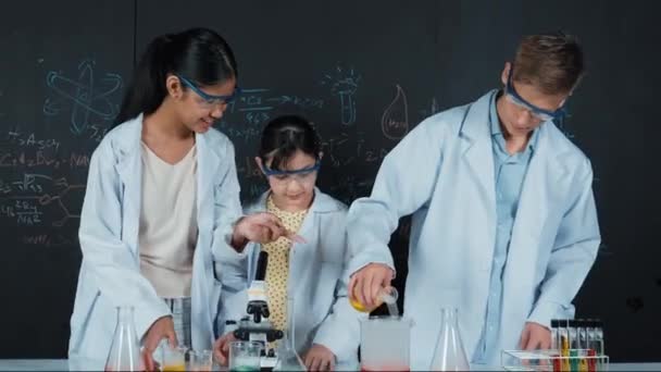 Kluger Junge mischt Flüssigkeit in Becher, während diverse Studenten experimentieren. Akademiker tragen Laborkittel, während sie farbige Proben mit Mikroskop und Versuchsausrüstung an den Tisch gießen. Erbauung - Filmmaterial, Video