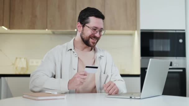 Happy male holding plastic card and feeling joy of large profit of money in own bank account indoors. Bearded man sitting in front of opened wireless laptop and looking on in with twinkle in eyes. - Footage, Video