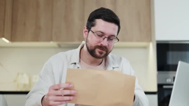 Shocked caucasian male open crafted envelope and taking out letter with bad news indoors. Surprised man with glasses feeling frustrated during reading important information at kitchen interior. - Footage, Video