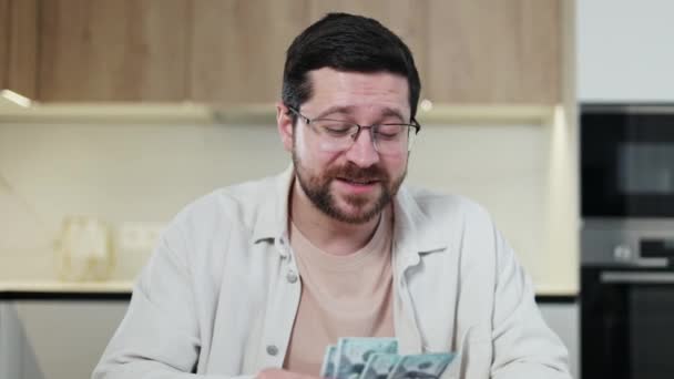 Uśmiechnięty nieogolony mężczyzna liczący banknoty dolarów i rozmawiający z kamerą z zadowoloną twarzą. Podekscytowany mężczyzna sprawdzanie kwoty zarobionych pieniędzy i uczucie zadowolenia z wyniku w mieszkaniu. - Materiał filmowy, wideo
