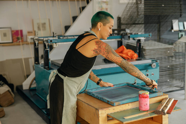 Naispuolinen työntekijä, jolla on squeegee, painaa mustetta runkoon, joka toimii design-studiossa. Silkkipaino - Valokuva, kuva
