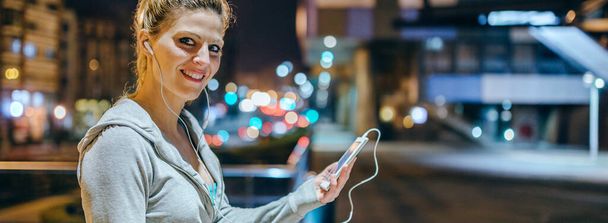 Portrait of smiling young blonde sportswoman listening music on mobile phone application. Banner of female runner with earphones looking at camera after training at night on town. Right copy space. - Photo, Image