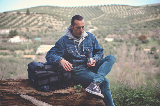 Backpacked ταξιδιώτη περιπετειώδη άνδρα κάθεται σε ένα κούτσουρο στα βουνά, πίνοντας τον πρωινό καφέ του από θερμός κύπελλο, χαλαρώνοντας στη φύση. Ανθρώπους. Υγιεινός τρόπος ζωής Δραστηριότητα αναψυχής Σαββατοκύριακου - Φωτογραφία, εικόνα