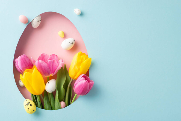 Szeszélyes húsvéti elrendezés felülnézetből, fényes tulipánokkal és festett tojásokkal, amelyek egy ovoid nyílásból kukucskálnak ki egy pasztell kék vásznon, helyet hagyva az üdvözlésre - Fotó, kép