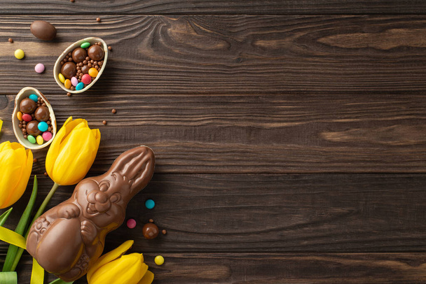 Easter delight perspective: top view photograph of chocolate egg broken apart to display assorted candies, alongside a chocolate hare and fresh tulips on a wooden table, with a blank area for text - Photo, Image