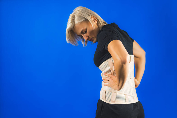 Woman in pain from back injury wearing lumbar brace corset on a blue background, copy space. High quality photo - Photo, Image