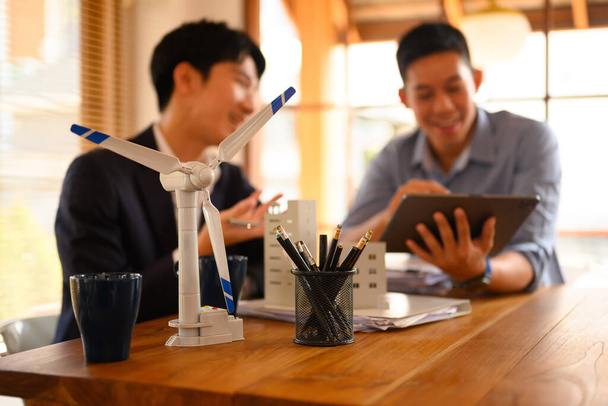 Un modèle d'éolienne sur table avec deux hommes d'affaires travaillant en arrière-plan. Concept d'énergie alternative. - Photo, image