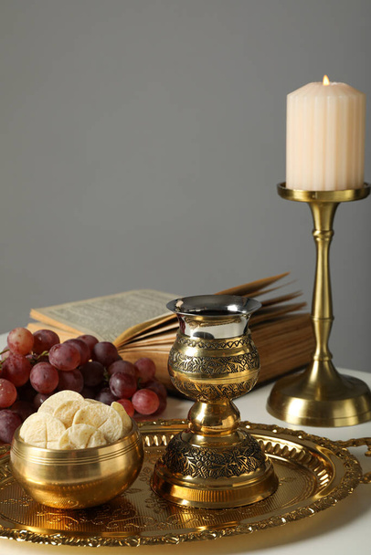 Свічка, книга, виноград, літургійний хліб, золотий хрест і чашка на столі на сірому фоні - Фото, зображення