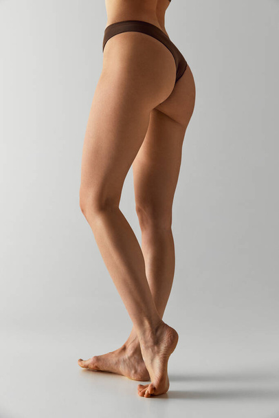 Glatte und durchtrainierte Beine. Zugeschnittenes Foto junger weiblicher Beine, Gesäß vor grauem Studiohintergrund. Model in Unterwäsche. Konzept der Schönheitsbehandlungen, Diäten, weibliche Gesundheit, Wellness-Verfahren. Anzeige - Foto, Bild