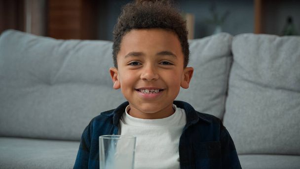 Kafa resmi, 10 'lu yaşlardaki komik, küçük, şirin, etnik çocuk evde süt içiyor çarpık dişleriyle kameraya gülümsüyor. Afro-Amerikan oğlu bir bardak tatlı organik yoğurt içiyor. - Fotoğraf, Görsel