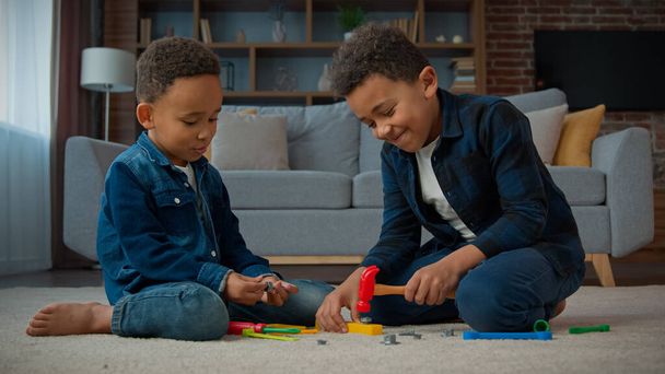 Δύο εθνοτικά μικρά αγόρια αφροαμερικανικά πολυφυλετικά παιδιά μαθητές παίζουν παιχνίδι κάθονται στο χαλί στο σαλόνι σε εσωτερικούς χώρους στο σπίτι αδελφοί φίλοι παίζουν με πλαστικά παιχνίδια εργαλεία επισκευής - Φωτογραφία, εικόνα