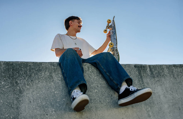Low angle of young male skater in casual outfit sitting on ramp with skateboard against blue sky - Photo, Image
