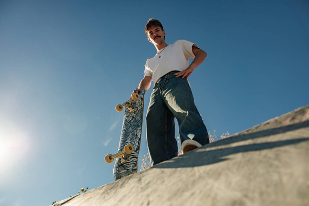 Low angle of happy male skateboarder holding skateboard standing at skate park in sunny day - Zdjęcie, obraz
