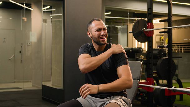 Latin kökenli çok ırklı Amerikalı güçlü bir adam spor salonunda oturuyor halterleri kaldırıyor ağır metal ekipmanları çalıştırıyor omuz ağrısı çekiyor erkek sporcu vücut geliştirme sporu sakatlığı - Fotoğraf, Görsel