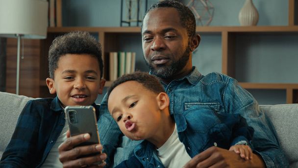 Afrikai amerikai család apa férfi és a kisgyerekek fiúk fiúk gyerekek ülni kanapén nézni mobil képernyőn szórakozás telefonnal próbálja vicces app fénykép maszk csinál szelfi grimasz látszó mobiltelefon otthon - Fotó, kép