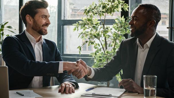 Two diverse multiracial businessmen signing document agreement shaking hands success business deal. Multiethnic men partners in office meeting handshake satisfied agree sign loan investment contract - Photo, Image