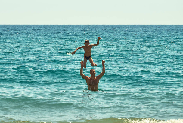 Glückliche Momente und Emotionen. Freudiger Vater hebt aufgeregten, glücklichen Sohn hoch. Wasser plätschert. Ruhe auf dem Meer. - Foto, Bild