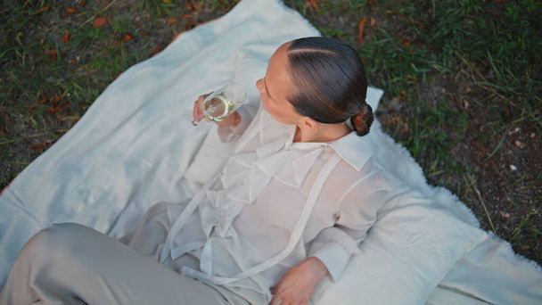 Pretty girl tasting wine on white blanket at picnic top view. Elegant woman drinking champagne from glass goblet lying on green grass. Relaxed beautiful lady closing eyes enjoying beverage on nature. - Photo, Image