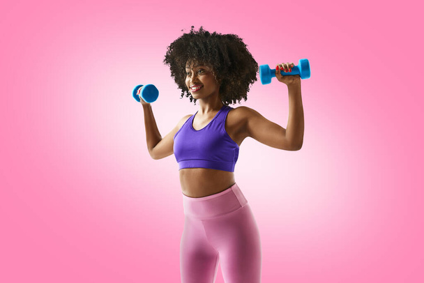 Smiling woman with dumbbells raised, wearing a purple sports bra and leggings against gradient pink studio background. Concept of sport, mourning routine, active and healthy lifestyle, energy, action. - Photo, Image