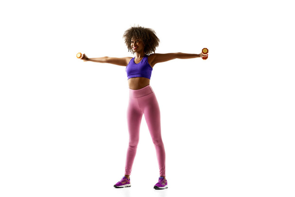 Fröhliche Afroamerikanerin, die mit blauen Gewichten trainiert, in lila Fitnessbekleidung vor weißem Studiohintergrund. Konzept von Sport, Trauerroutine, aktiver und gesunder Lebensweise, Energie. - Foto, Bild