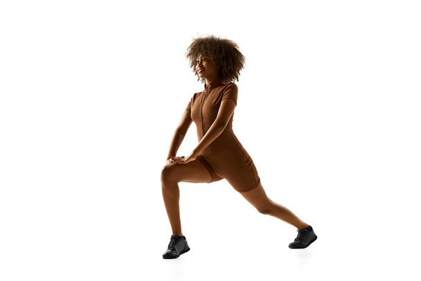 Fitness instructor demonstrating lunge training in brown activewear against white studio white background. Exercises for legs. Concept of sport, mourning routine, active and healthy lifestyle, action. - Foto, Imagen
