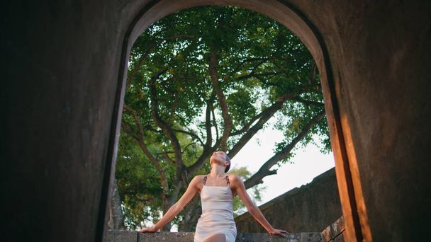 Gorgeous girl standing palace in elegant white dress view from arch. Beautiful young woman posing at castle balcony in front green trees. Relaxed sensual model looking camera leaning on stone fence. - Photo, Image