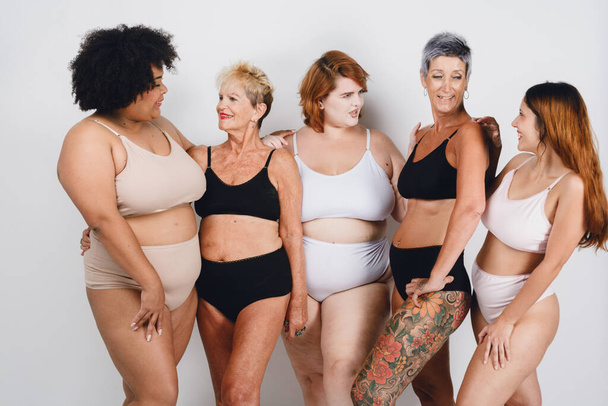 5 mulheres de diferentes etnias e idades posando, afro, caucasiana, morena, gorda e magra, com cabelo curto e longo, estão em roupa interior em fundo branco olhando umas para as outras - Foto, Imagem