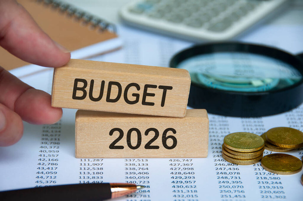 テキストで木製のブロックを保持する手 - 財務データの背景を持つ予算2026. 予算コンセプト - 写真・画像