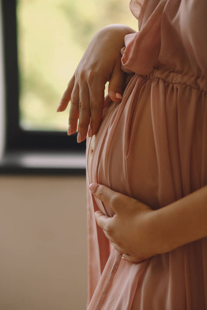 Pembe elbiseli hamile bir kadın pencerenin yanında duruyor ve içeride karnına dokunuyor. Hamile karnının yakın fotoğrafını çek. Tarz sahibi annelik kavramı - Fotoğraf, Görsel