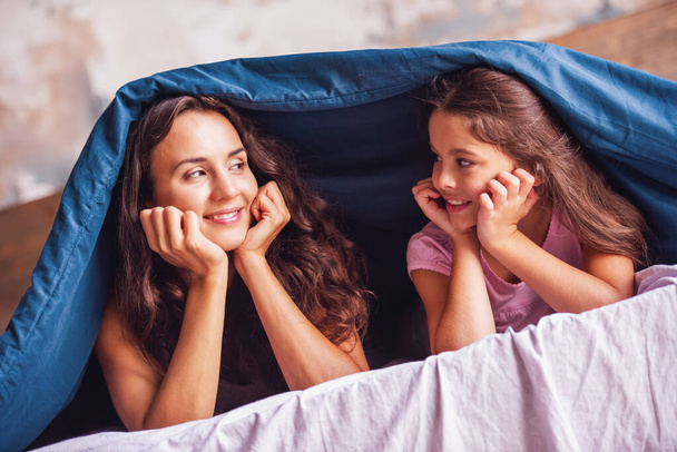 Kaunis äiti ja tytär katsovat toisiaan ja hymyilevät sängyssä peitettynä pussilakanalla.
 - Valokuva, kuva
