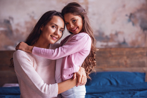 Bela mãe e filha estão abraçando, olhando para a câmera e sorrindo enquanto estão sentados na cama em casa
 - Foto, Imagem