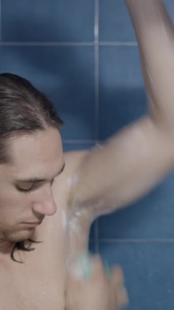 若い慎重な長髪の男は,彼の脇の下をこすり,シャワーで彼の腕 - クローズアップショット - 垂直ビデオ - 映像、動画