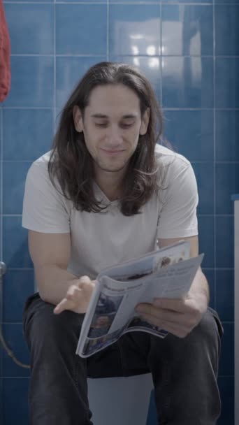 Young caucasian man reading a newsletter in the bathroom while shit - Frontal shot - Vertical video - 映像、動画