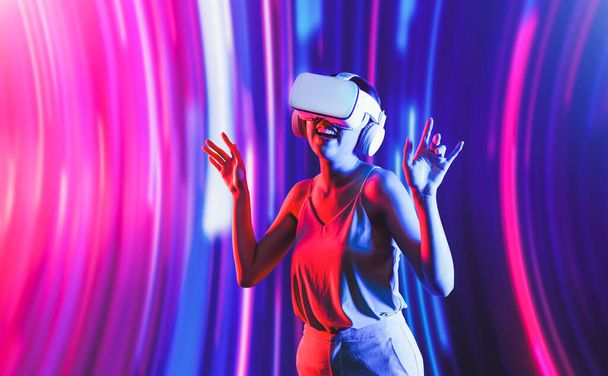 Älykäs nainen seisoo neonvalon ympäröimänä käyttäen VR-kuulokkeita, jotka yhdistävät metaversen, tulevan kyberavaruusyhteisön teknologian. Tyylikäs nainen nauttii tanssia horjuva elin ja laulaa laulun. Aistiharhat. - Valokuva, kuva