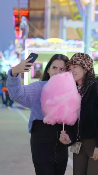 Jóvenes amigas se toman selfies haciendo caras graciosas sosteniendo algodón de azúcar en el parque de atracciones por la noche en Valencia - FullHD Vertical video - Imágenes, Vídeo