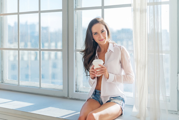 Bella ragazza che indossa abiti sexy casa seduta accanto alla finestra mattina con una tazza di caffè nelle sue mani piene di luce
 - Foto, immagini