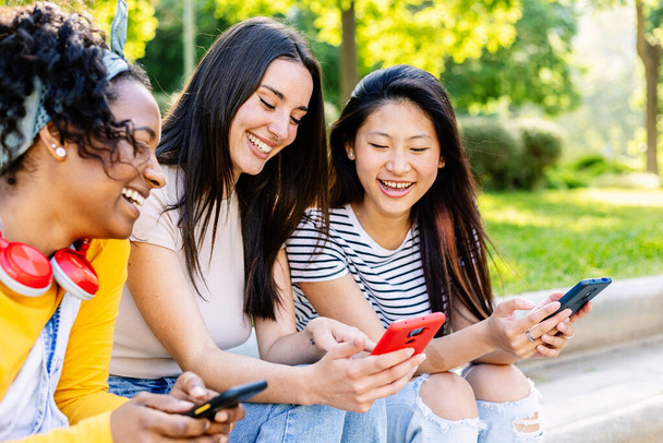 Tre giovani donne che utilizzano telefoni cellulari sedute insieme al parco cittadino in una giornata di sole. Stile di vita tecnologico e concetto di social media. - Foto, immagini