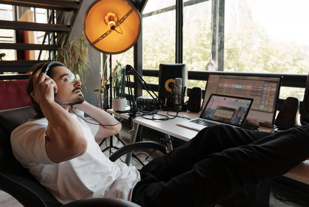 Musicien masculin pensif en vêtements décontractés et écouteurs assis près d'un ordinateur portable avec les jambes sur la table tout en composant de la musique et podcasting en studio d'enregistrement - Photo, image