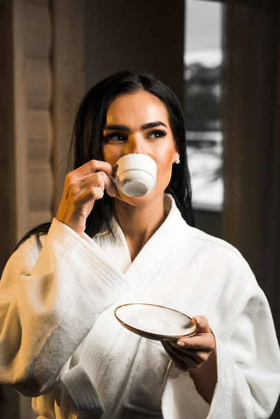 Молодая женщина в белом халате пьет кофе эспрессо в деревянном номере отеля. Концепция отдыха в отеле. Утренний кофе - Фото, изображение