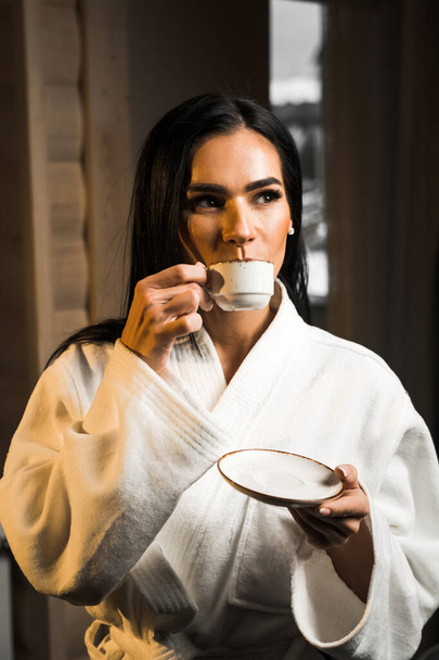 Νεαρή γυναίκα με λευκό μπουρνούζι πίνει καφέ εσπρέσο σε ξύλινο δωμάτιο ξενοδοχείου. Ξενοδοχείο έννοια διακοπών. Πρωινός καφές - Φωτογραφία, εικόνα