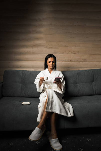 Uma jovem de roupão branco bebe café expresso num quarto de hotel de madeira. Conceito de férias de hotel. Café da manhã - Foto, Imagem