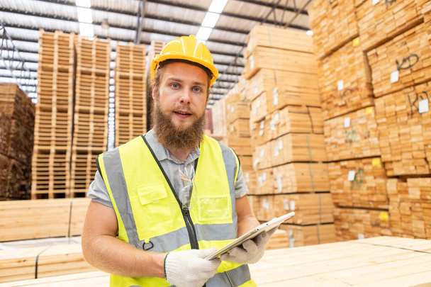 Munkás Férfi szakállas visel biztonsági egyenruha sárga kemény kalap segítségével tabletta dolgozik ellenőrzése minőségű fa gyártása fa. Férfi asztalos munkás faipari raktár ipar. - Fotó, kép