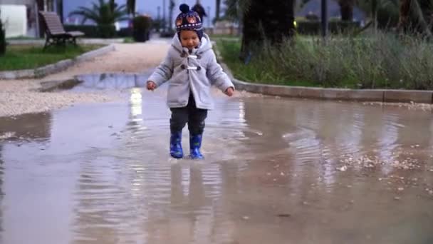 Little girl in a jacket and rubber boots stomping on a puddle in the park. High quality 4k footage - Footage, Video