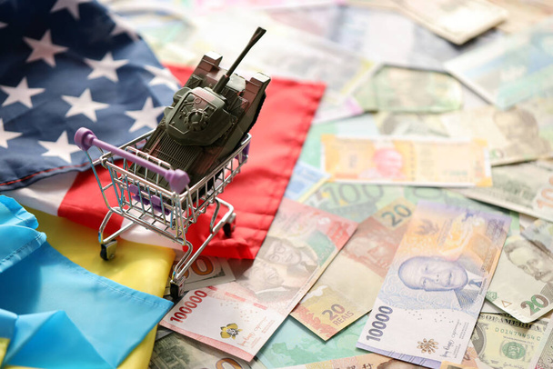 Zbiornik zabawek na fladze amerykańskiej i fladze ukraińskiej na wielu banknotach różnych walut. Kontekst finansowania działań wojennych i wsparcia wojskowego między Stanami Zjednoczonymi a Ukrainą - Zdjęcie, obraz