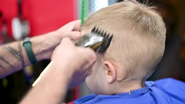 Közelről láthatjuk a fodrász fiúk fejét. Egy hozzáértő borbély elektromos borotvát használ a fodrászatban. Kis ügyfél egyre stílusos férfi frizura. - Felvétel, videó